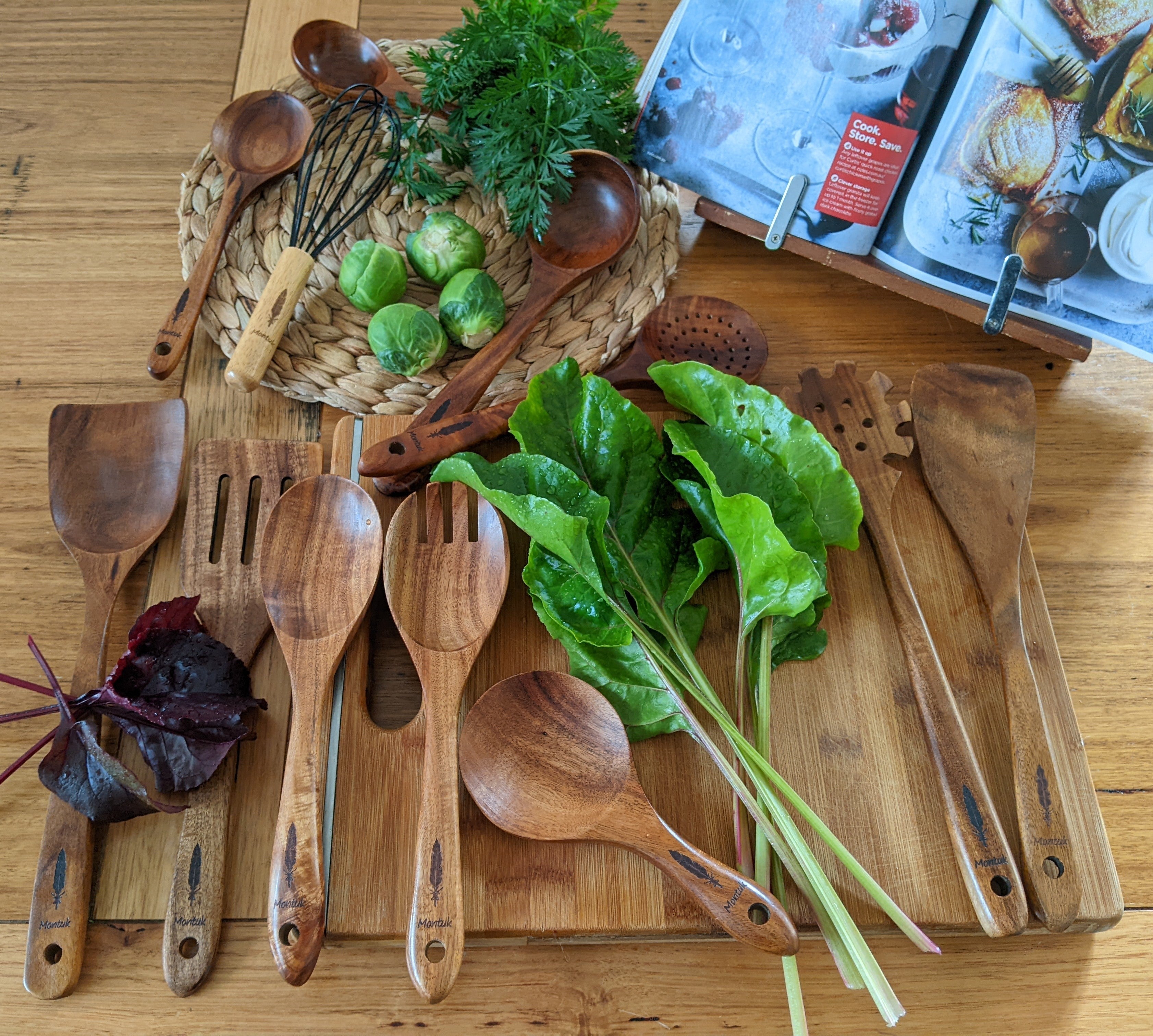 Teak wood cooking utensil set styled on a wooden board with fresh greens